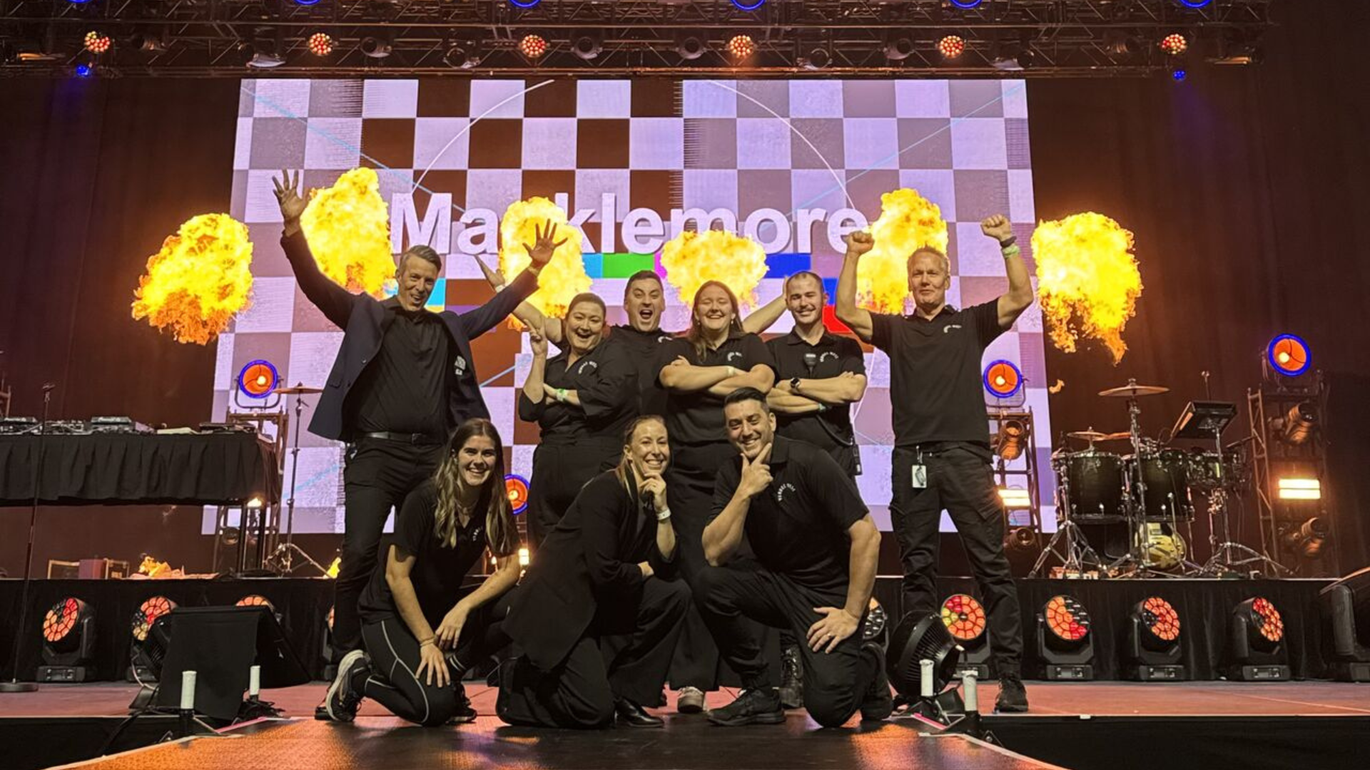 A photo of VenuesWest employees posing on a stage in front of a "Macklemore" sign